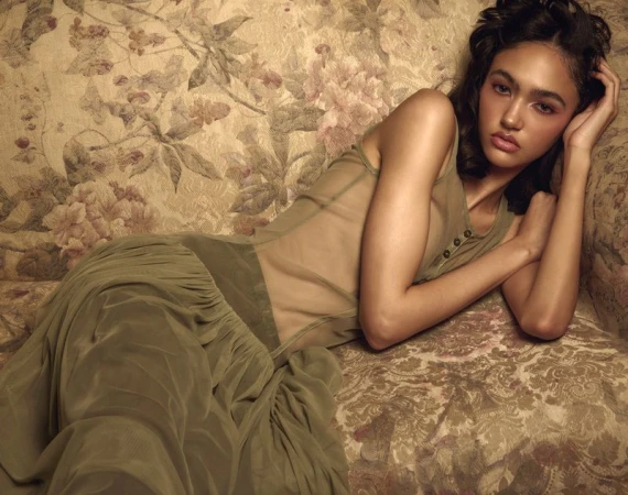 Model Jacki Girard reclining on a vintage floral upholstered sofa wearing a sheer olive green dress.