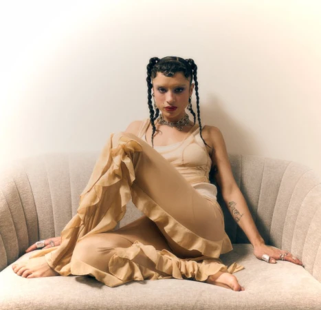 Woman with braided hair in a ruffled beige outfit and statement jewelry sitting barefoot on a curved fluted sofa.