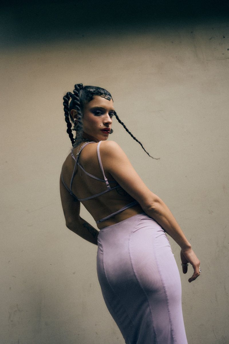 Woman in a lilac backless strappy top and matching skirt looking over her shoulder against a neutral wall.