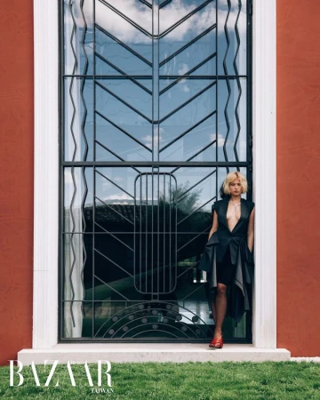 Model Ksenia Lifanova in a black asymmetrical dress poses beside a tall geometric iron window set in a terracotta exterior wall.