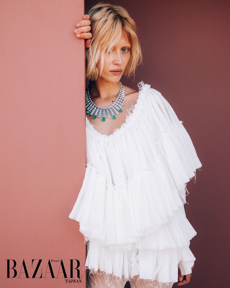Model Ksenia Lifanova wearing an emerald statement necklace and a tiered white ruffled dress leaning against a peach wall.