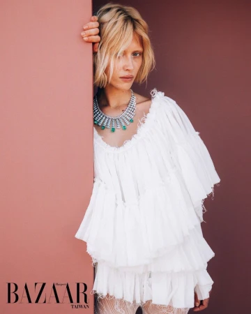 Model Ksenia Lifanova wearing an emerald statement necklace and a tiered white ruffled dress leaning against a peach wall.