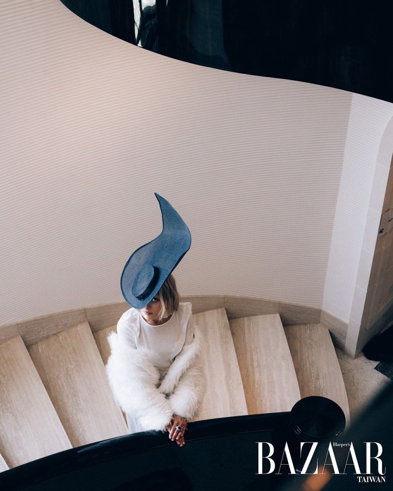 High-angle shot of a woman wearing a sculptural blue crescent hat and a white fur coat on a stone spiral staircase.