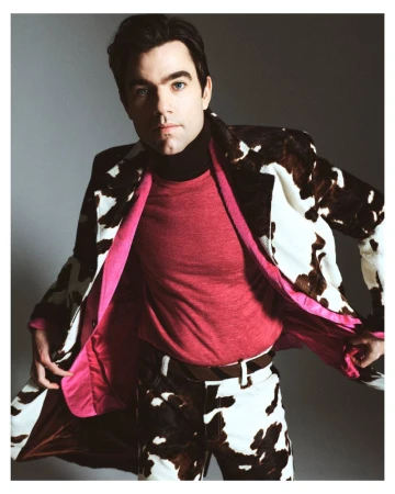 Man in a cow-print coat with vibrant pink lining and a pink top posing against a studio backdrop.