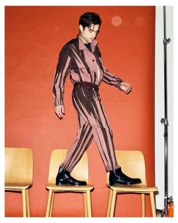 Patrick Ball wearing a patterned two-piece suit and black boots while balancing across wooden studio chairs against an orange backdrop.