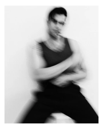 Monochrome motion-blurred portrait of a man wearing a dark sleeveless tank top and trousers against a white seamless background.