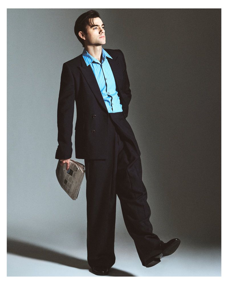 Patrick Ball in an oversized black suit and light blue shirt holding a newspaper against a grey studio backdrop.