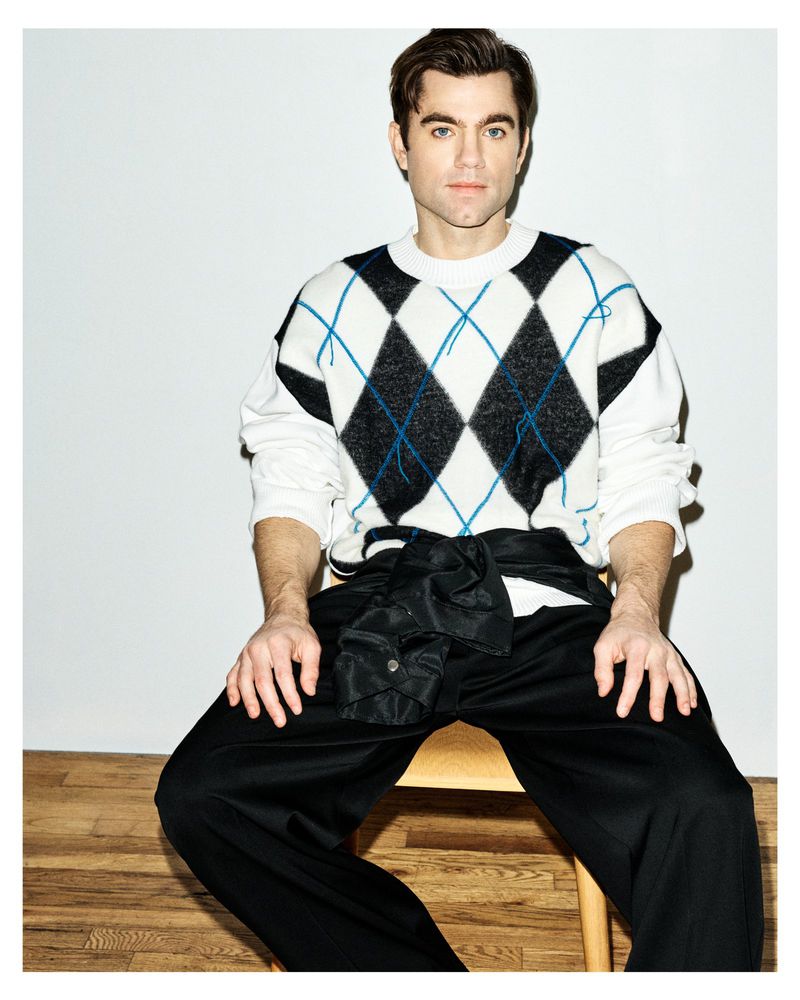 Patrick Ball seated on a chair wearing a white and black argyle knit sweater with a jacket tied around his waist.