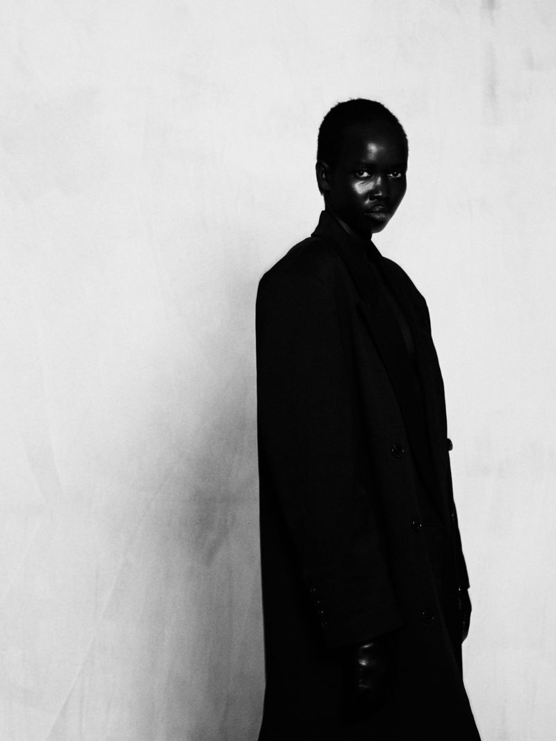 Black and white portrait of Akuel Malong wearing a black tailored coat against a textured pale studio background.