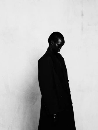 Model Akuel Malong looking down wearing a structured long black coat against a textured pale studio wall.