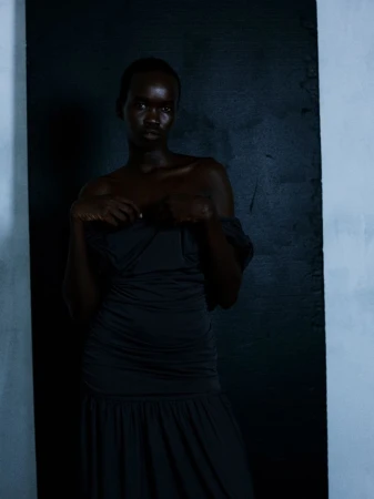 Model Akuel Malong wearing a dark off-the-shoulder ruched dress in low-key lighting against a dark studio backdrop.