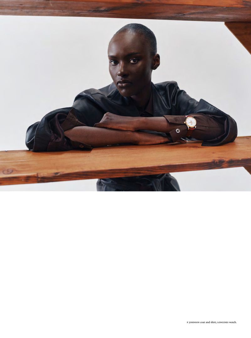 Model Amou Ring leaning on a wooden shelf wearing a dark coat and wristwatch against a white studio background.