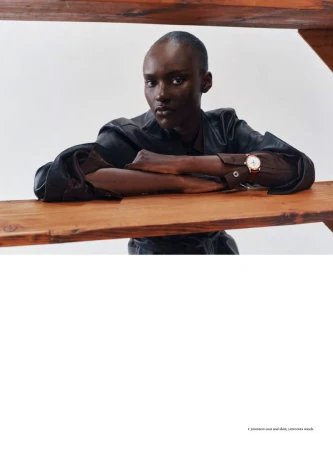 Model Amou Ring leaning on a wooden shelf wearing a dark coat and wristwatch against a white studio background.