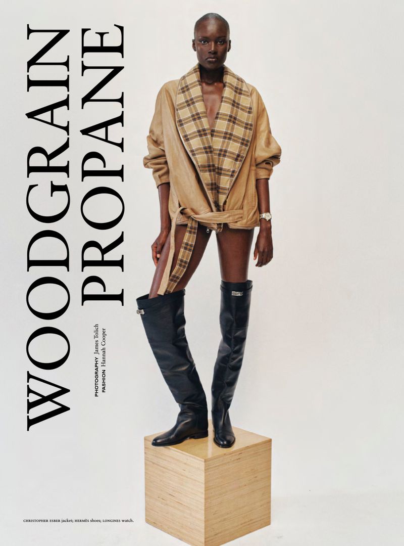Model Amou Ring standing on a wooden plinth in a camel belted check-lapel jacket and black over-the-knee leather boots.