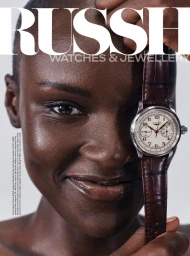 Model Amou Ring holding a luxury watch with a brown leather strap beside her face for Russh magazine on a white background.