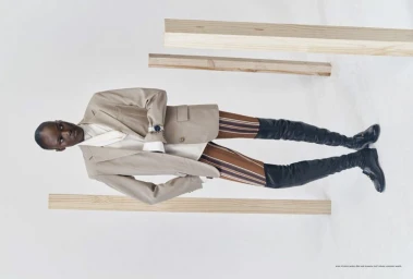 Model Amou Ring poses in a tailored beige blazer, striped tights, and tall black boots among wooden planks in a white studio.