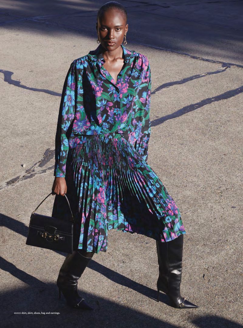 Model Amou Ring in a floral pleated skirt and shirt set with black leather knee-high boots holding a black handbag on pavement.