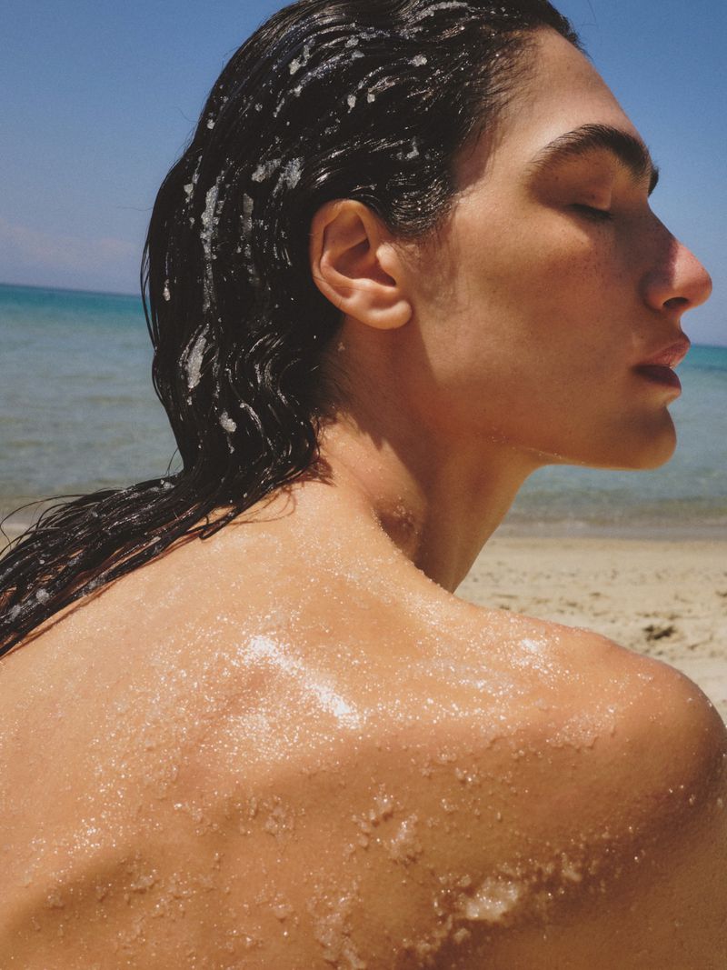 Close-up profile of a woman with wet dark hair and glistening sea salt crystals on her bare shoulder and back at the beach.