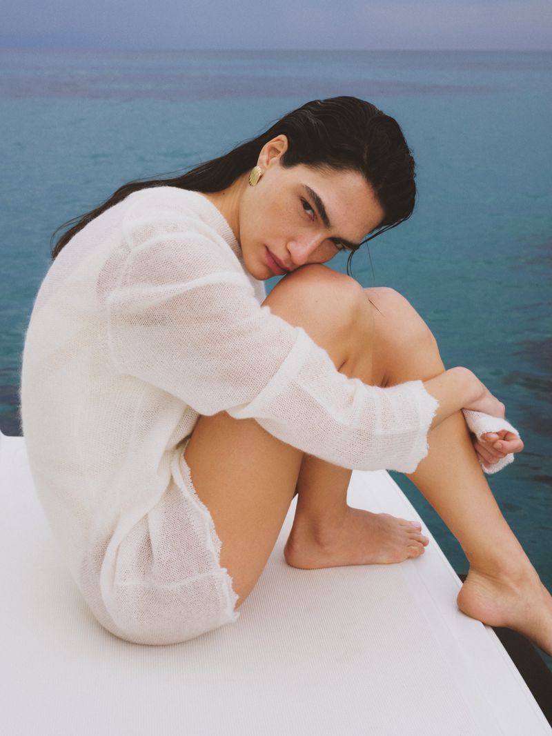 Model with slicked back dark hair wearing a sheer white knit sweater seated barefoot on a boat deck overlooking calm blue ocean water.