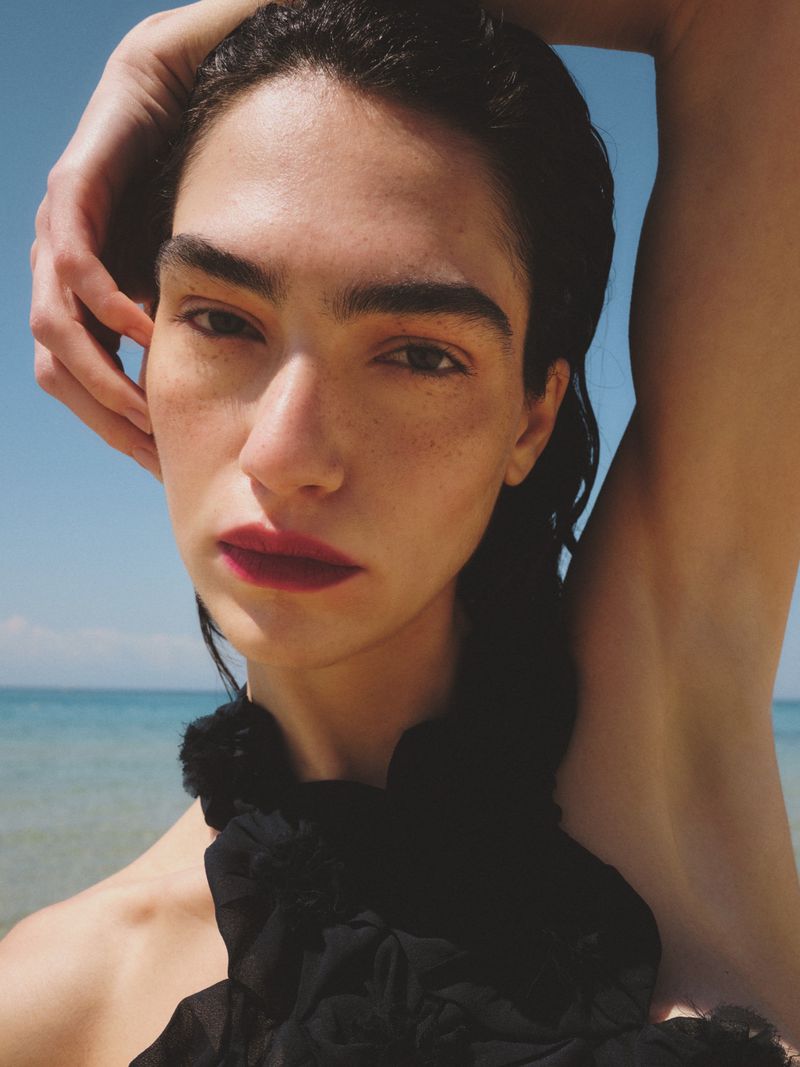 Close-up portrait of a woman with slicked dark hair and bold lipstick posing with an arm raised against a calm ocean backdrop.