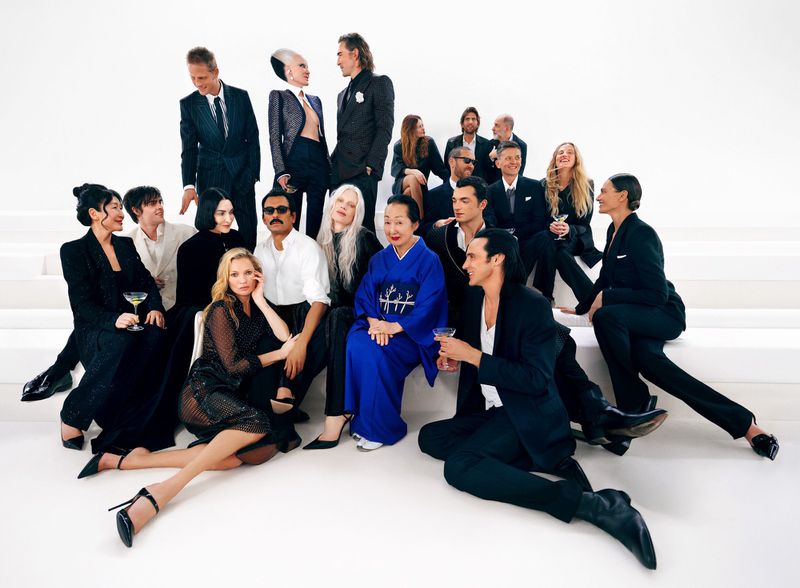 A large ensemble of models and celebrities in black formal tailoring posing together against a bright white seamless studio background.