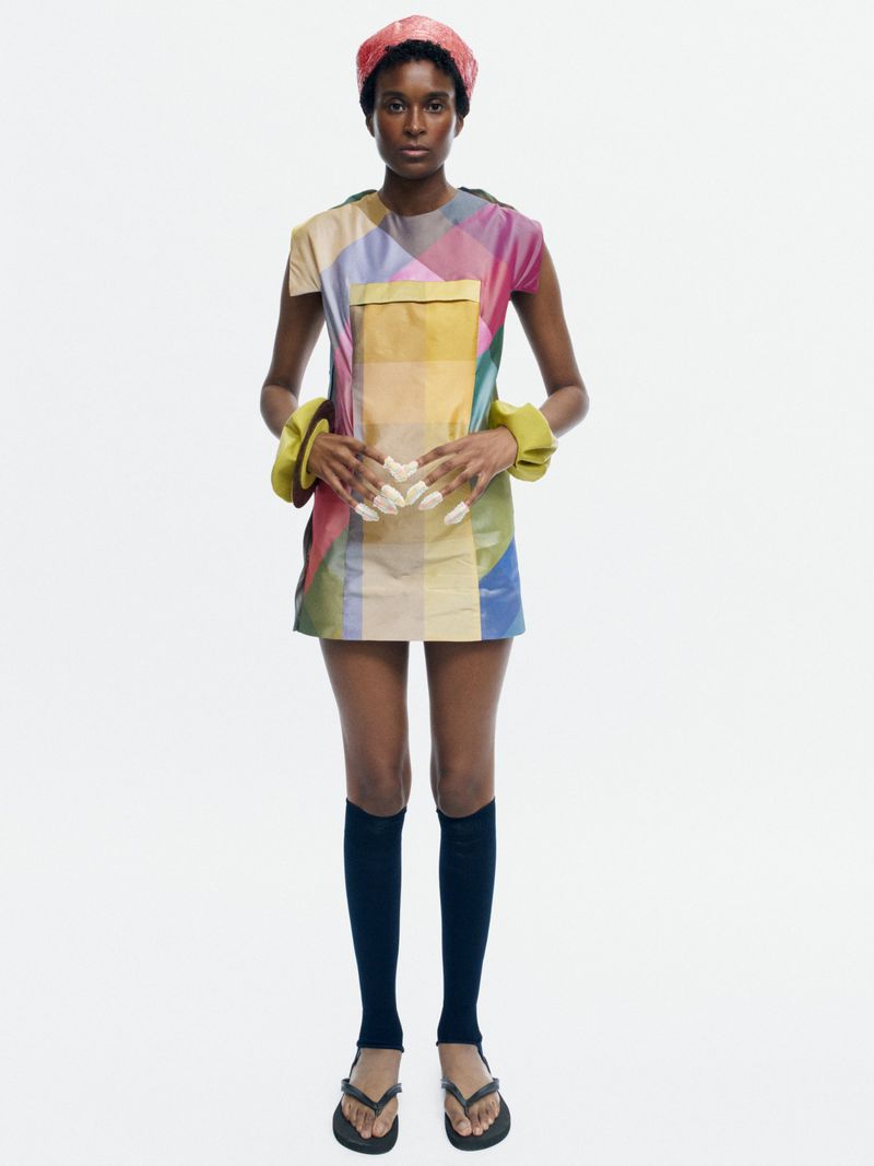 Model Bella Berghoef standing in a multicolor geometric shift mini dress, pink cap, and black knee-high socks on a white background.