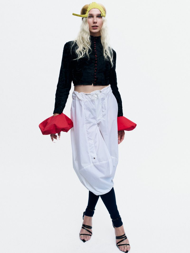 Model Bella Berghoef wearing a cropped black jacket, oversized red cuff accents, and white draped trousers on a white backdrop.