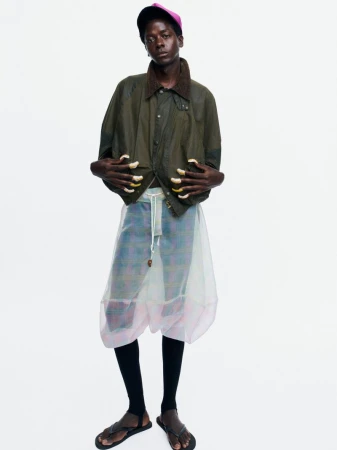 Black male model in an olive green jacket, sheer layered pants, and a pink cap standing against a white seamless backdrop.