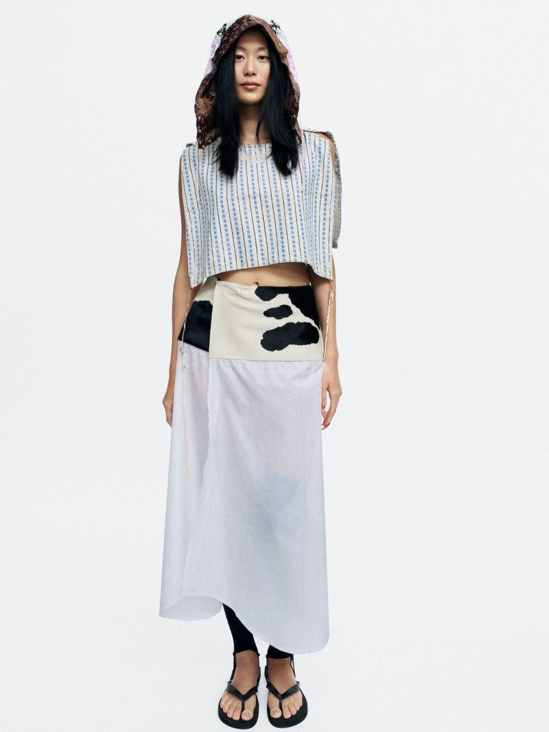 Model Bella Berghoef standing in a cropped sleeveless top, white wrap skirt with black pattern, and hooded headpiece on white background.