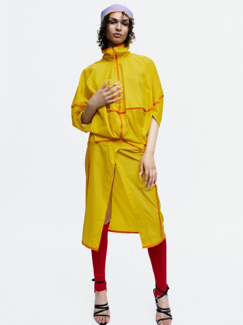 Bella Berghoef wearing a bright yellow nylon two-piece outfit and red knee socks against a white studio background.