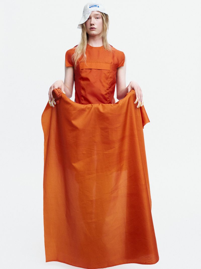 Model Bella Berghoef wearing a white bucket hat and holding a draped bright orange garment against a white seamless background.