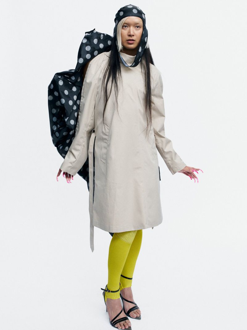 Model Bella Berghoef in a beige coat, polka dot headpiece, yellow tights, and strappy heels against a white studio background.