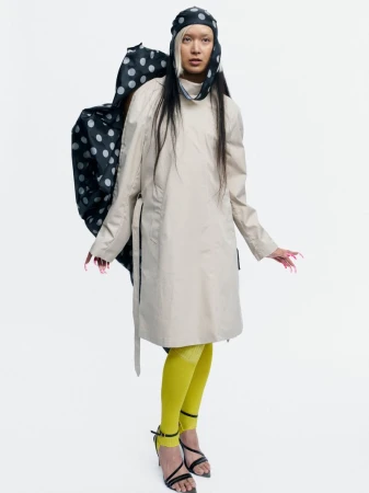 Model Bella Berghoef in a beige coat, polka dot headpiece, yellow tights, and strappy heels against a white studio background.