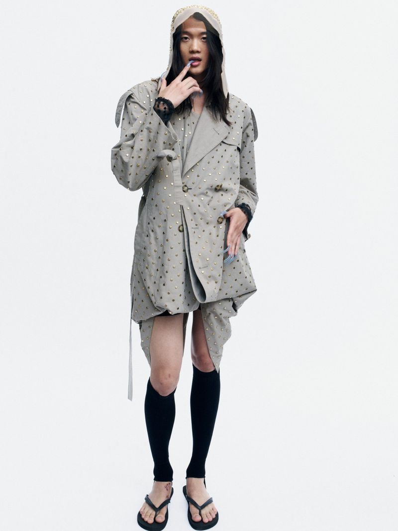 Model wearing an oversized studded beige trench coat, hood, black knee-high socks, and thong sandals against a white studio background.