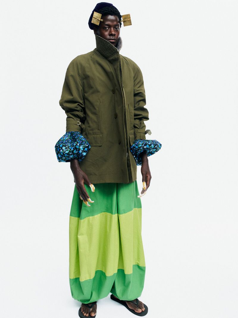 Full-length shot of a Black male model in an olive jacket and voluminous green colorblock trousers on a white seamless backdrop.