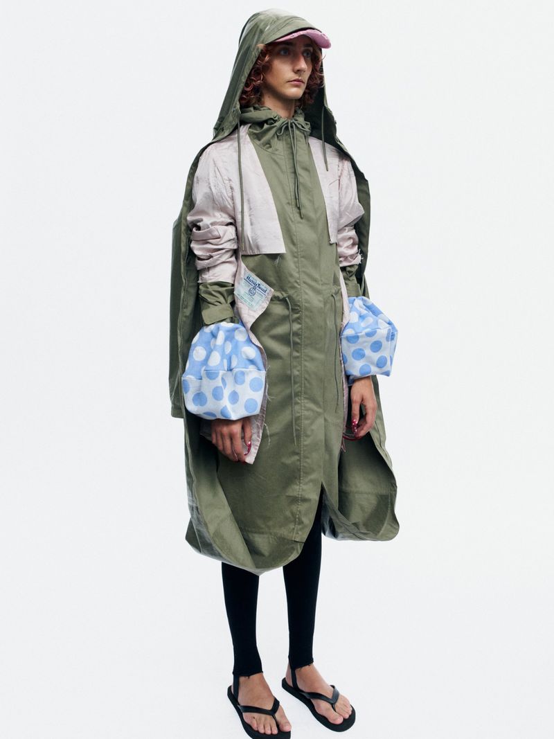 Model Bella Berghoef wearing an oversized olive green hooded parka with blue polka dot cuffs against a white seamless studio backdrop.