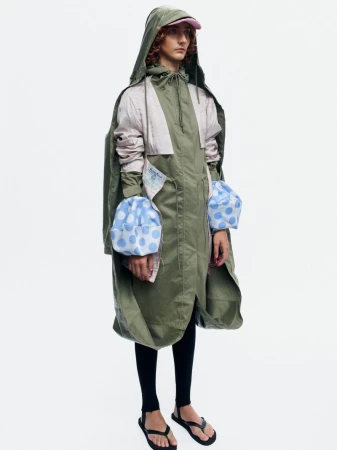 Model Bella Berghoef wearing an oversized olive green hooded parka with blue polka dot cuffs against a white seamless studio backdrop.