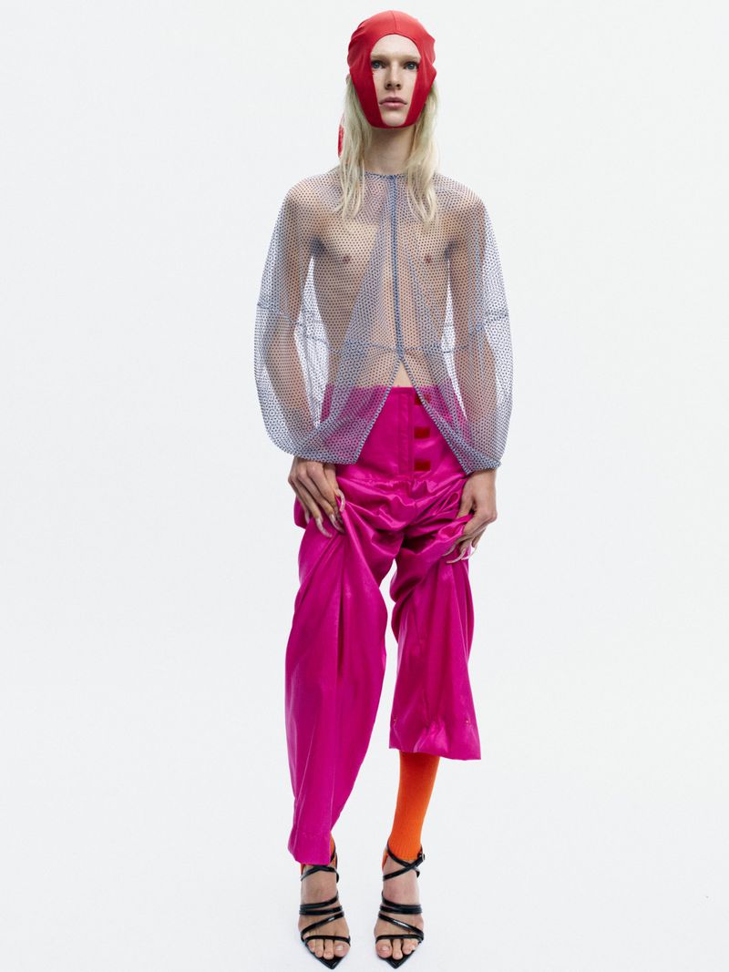 Model Bella Berghoef in a sheer mesh top, magenta satin trousers, and a red headpiece against a white seamless backdrop.