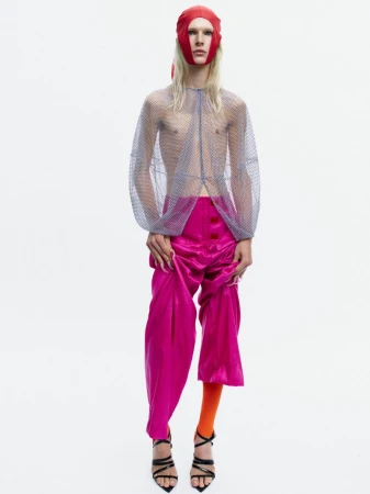 Model Bella Berghoef in a sheer mesh top, magenta satin trousers, and a red headpiece against a white seamless backdrop.