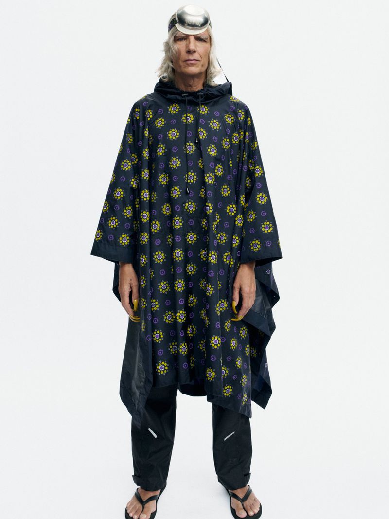 Model wearing a dark printed hooded poncho, black pants, and a metallic dome headpiece standing against a white seamless backdrop.