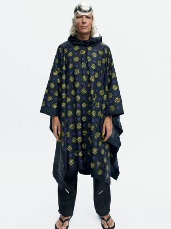 Model wearing a dark printed hooded poncho, black pants, and a metallic dome headpiece standing against a white seamless backdrop.