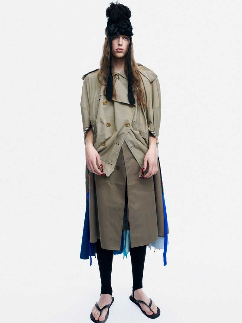Model Bella Berghoef wearing a layered beige trench coat, tall black headpiece, and black sandals against a plain white studio backdrop.
