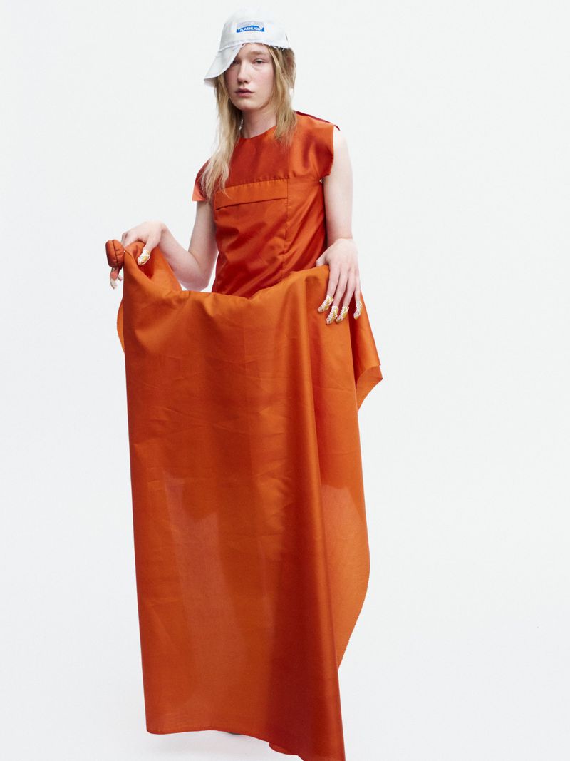 Model Bella Berghoef in an orange sleeveless top and white cap holding draped orange fabric against a white seamless background.