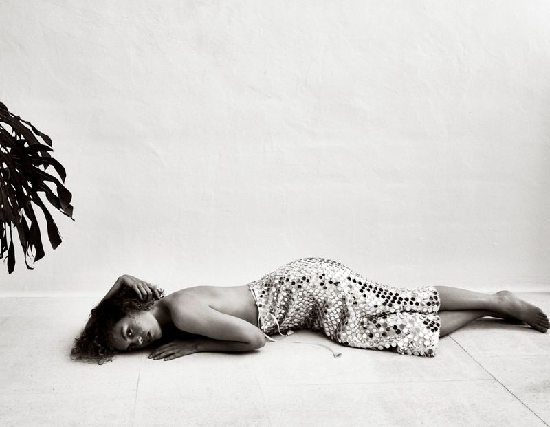 Monochrome portrait of a young woman lying on a tiled floor, wearing a polka dot dress, with a monstera leaf in the background.