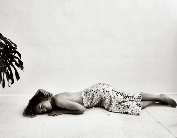 Monochrome portrait of a young woman lying on a tiled floor, wearing a polka dot dress, with a monstera leaf in the background.