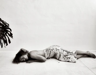 Model lying on the floor wearing a reflective sequined skirt against a plain wall with a tropical leaf shadow.