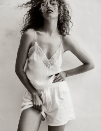 Model with curly hair wearing a white lace-trimmed camisole and white shorts against a textured background.