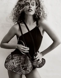 Monochrome fashion portrait of a curly-haired woman holding mesh bags against a plain wall