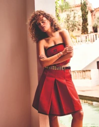 Model with curly hair wearing a red strapless top and matching red skirt posing outdoors near a pool.