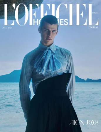 Male model in a light blue striped shirt with ruffled ascot and black structured skirt standing against a calm coastal sea.